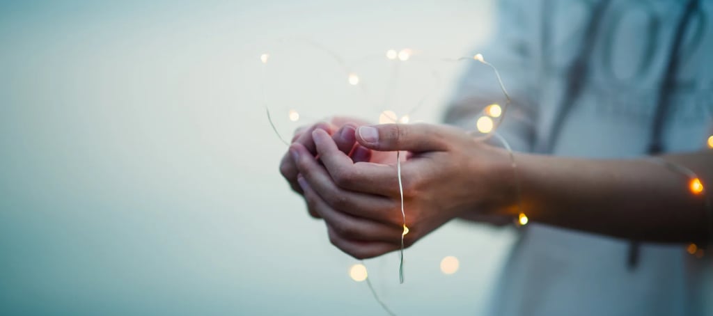Cupped hands holding warm glowing fairy lights in the shape of a heart against a soft blue background.