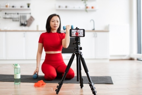 A fitness influencer filming a dumbbell arm workout at home using a smartphone on a tripod.