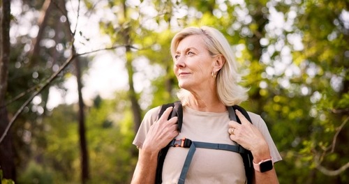 An active senior woman hiking in a sunlit forest with a backpack and smartwatch.