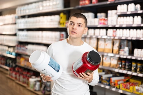 A man in a sports nutrition store comparing two large tubs of protein powder supplements.