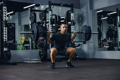 A fit man performing a heavy barbell back squat in a modern gym with weights and racks.