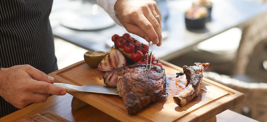 A chef slicing a grilled ribeye steak on a wooden cutting board with roasted vegetables.