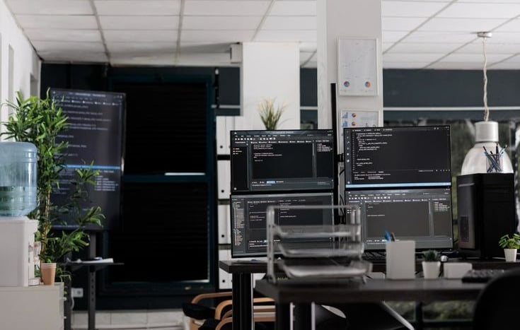 Kantor pengembangan perangkat lunak modern dengan beberapa monitor yang menampilkan kode, melambangkan lingkungan kerja softw