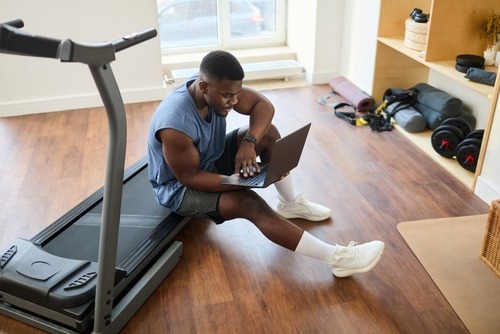 A man in gym clothes sits on a treadmill using a laptop for his home workout routine.