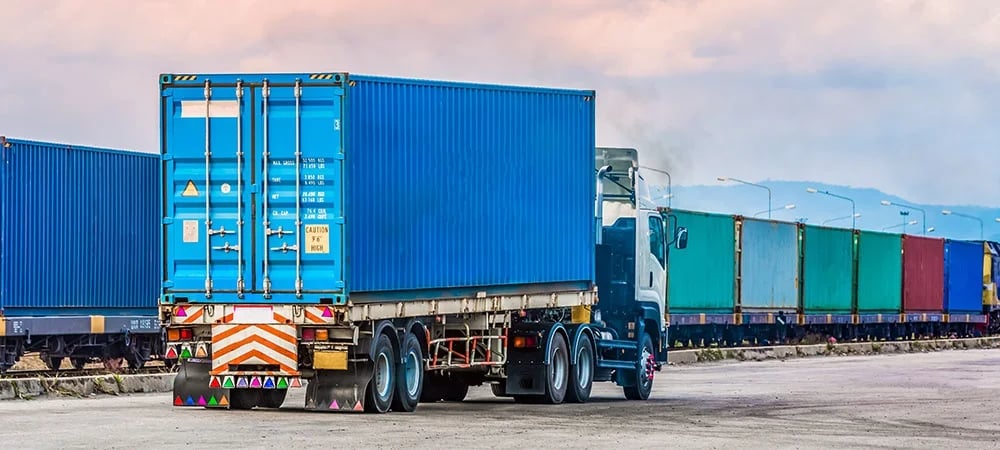 A logistics semi-truck carrying a blue shipping container near a freight train for intermodal transport.