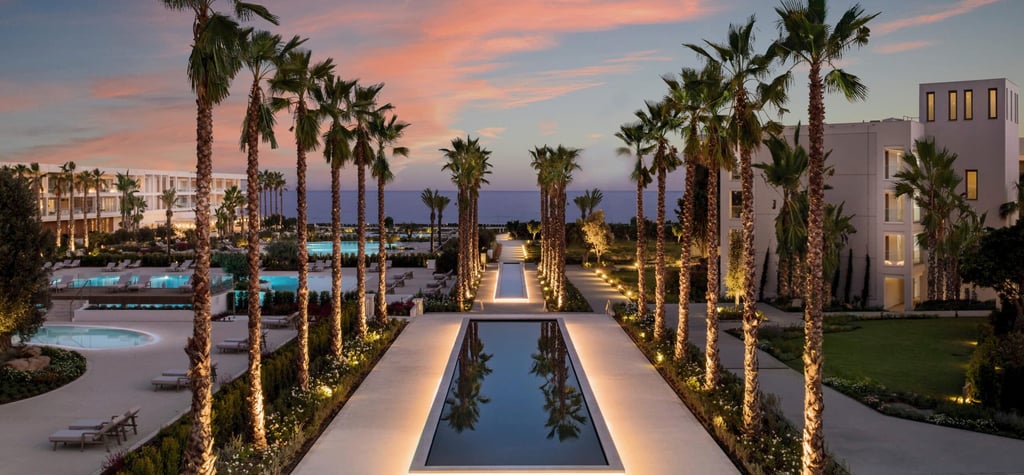 Luxury tropical resort at sunset with lit reflecting pools, palm trees, and ocean views.