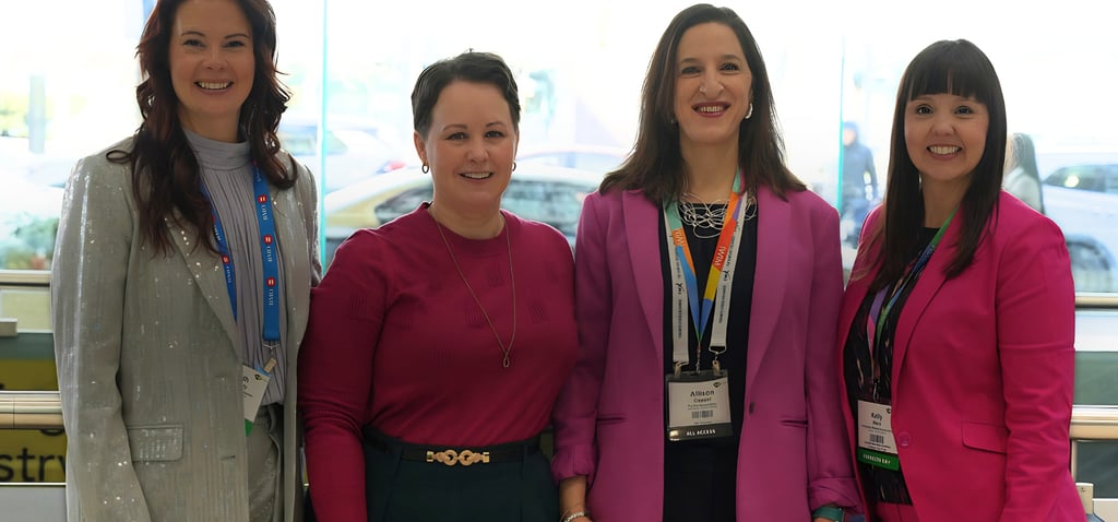 Four professional women standing by a window with a city view, wearing name tag lanyards and smiling for a group photo.