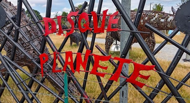 Red metal letters spelling Disque Planete on a black lattice fence near metal scrap sculptures.