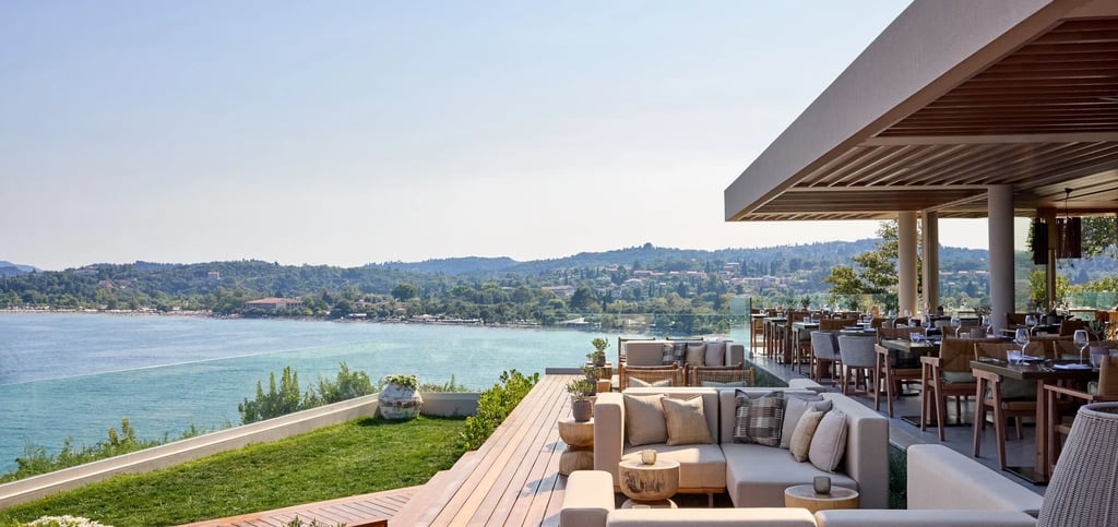 Ikos Odisia lounge and restaurant terrace overlooking a blue ocean bay and lush coastline.