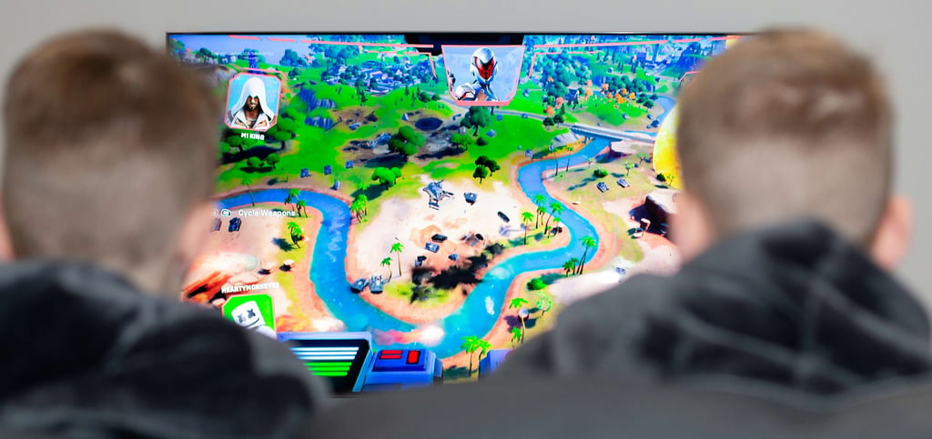 Two people playing the Fortnite video game on a large TV screen featuring a map with islands and rivers.