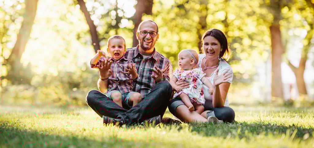 homem e mulher sentados em um gramado, cada um segura uma criança, expressão de felicidade no rosto