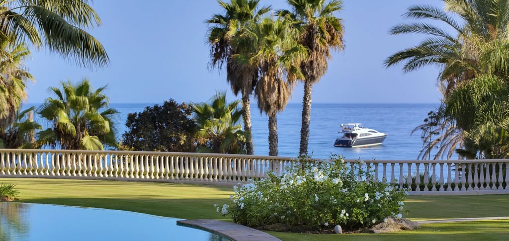 Luxury villa in Marbella overlooking a yacht on the blue ocean with palm trees.