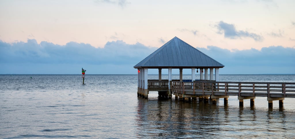 A wooden pier with a gazebo pavilion overlooks a calm ocean bay at sunrise with blue and pink clouds.