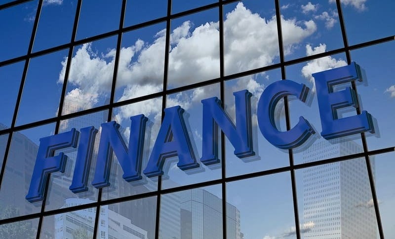 3D-Finanz-Schild an einem Glas-Bürogebäude mit Spiegelungen von weißen Wolken und blauem Himmel.