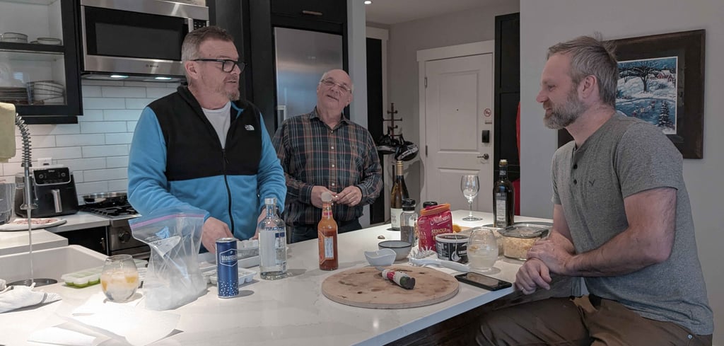 Three men talking around a kitchen island with drinks and food during a casual evening at home.