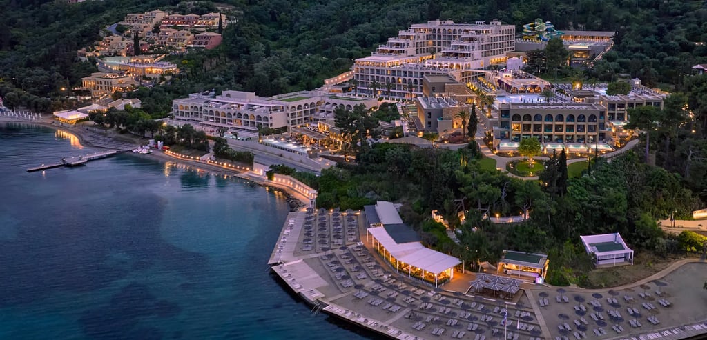 Aerial view of a luxury beachfront resort in Corfu at dusk with lighted pools and private beach.