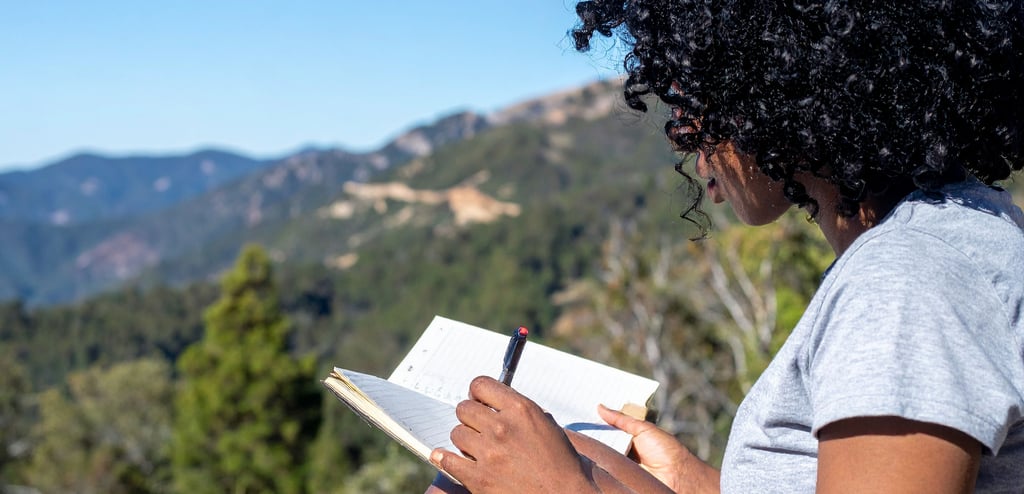A woman with curly black hair writes in her journal while sitting outdoors with a mountain view.
