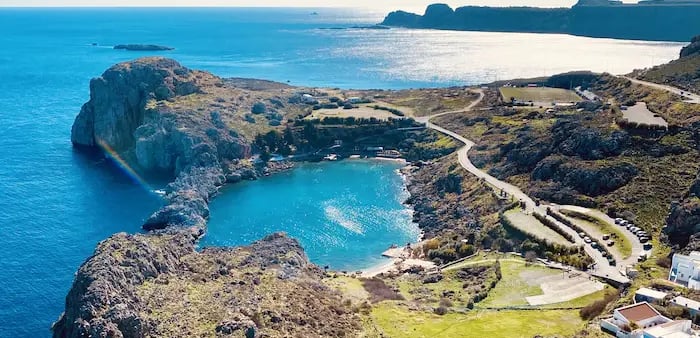Aerial view of St: Paul's Bay in Lindos, Rhodes, featuring turquoise waters and rocky Greek coastline.