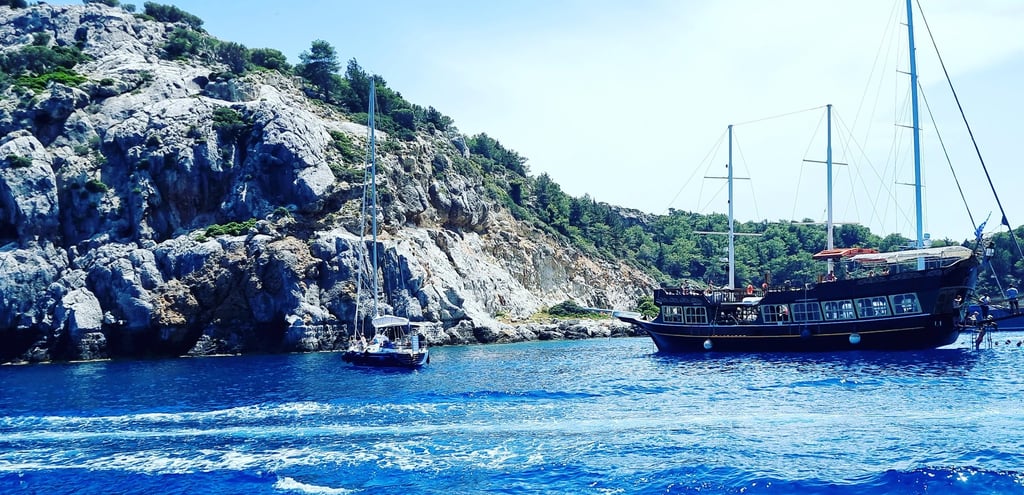 A wooden pirate ship and a sailboat anchor in the bright blue water of a Mediterranean bay.