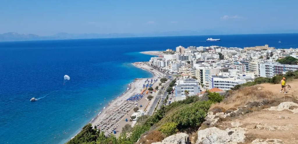 Aerial view of Rhodes City beach with white buildings, blue Aegean Sea, and parasailing on a sunny day.