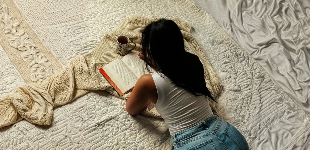 A woman relaxes on a cozy bed with a book, coffee, and a textured knit blanket.