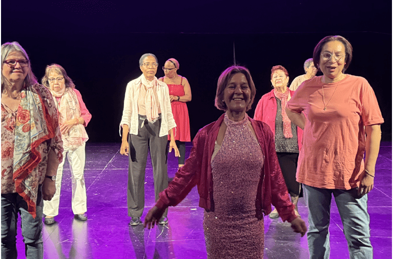 Diverse group of senior women performing a dance routine on a theater stage with purple lighting.