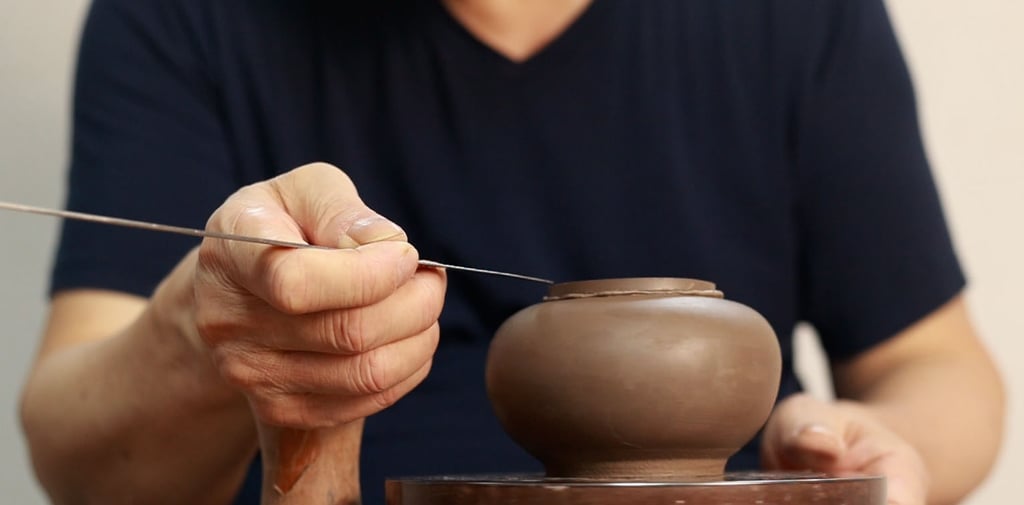 A man holding a long-handled pointed knife, carving the edge of a Yixing teapot