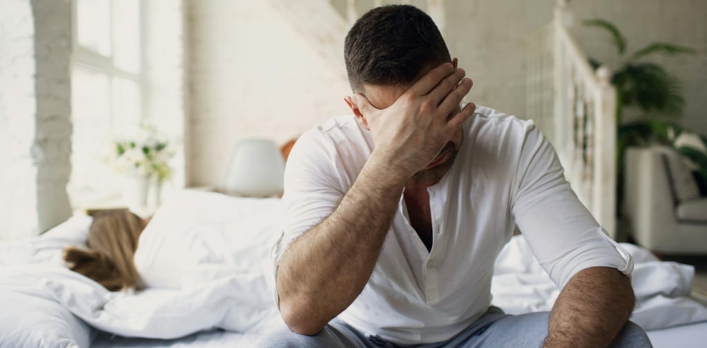Stressed man sitting on bed edge with head in hand while wife sleeps, depicting relationship problems and insomnia.