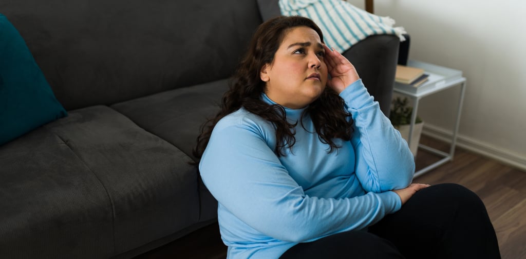 A concerned woman sitting on the floor by a sofa, holding her head to represent stress or a headache.