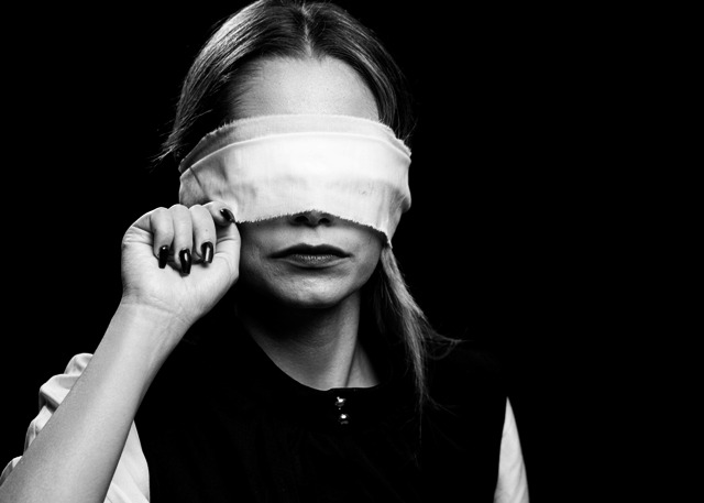 Kobieta z opaską na oczach w czarno-białym ujęciu, unosząca ją delikatnie palcami w kierunku światła