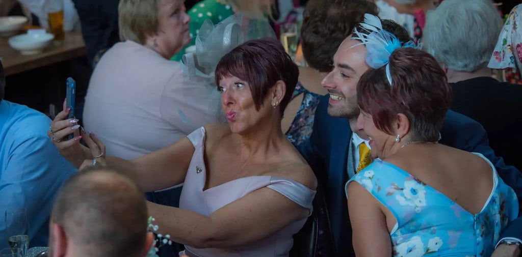 Wedding guests in elegant formal attire smiling and taking a group selfie with a smartphone at a reception table.