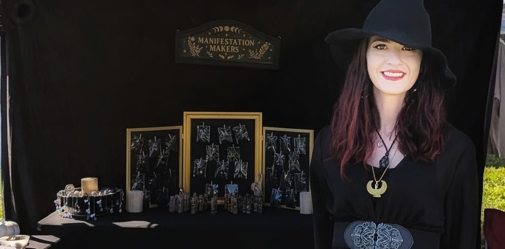 Woman in a black dress and witch hat in front of a Manifestation Makers sign and her jewelry display