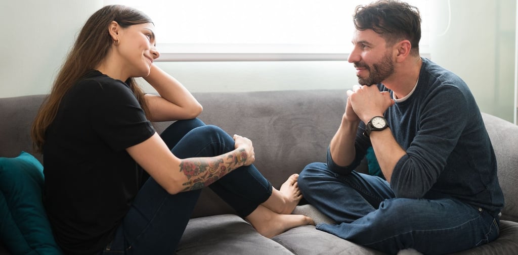 Moteris ir vyras sėdi ant minkštos sofos ir šypsosi žiūrėdami vienas į kitą.