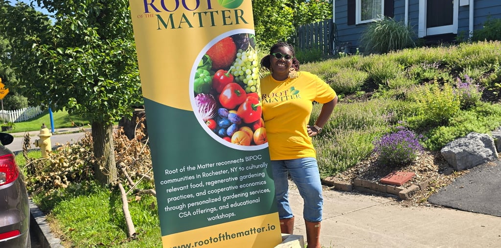 A woman in a yellow Root of the Matter t-shirt stands by a tall banner for community gardening in Rochester, NY.