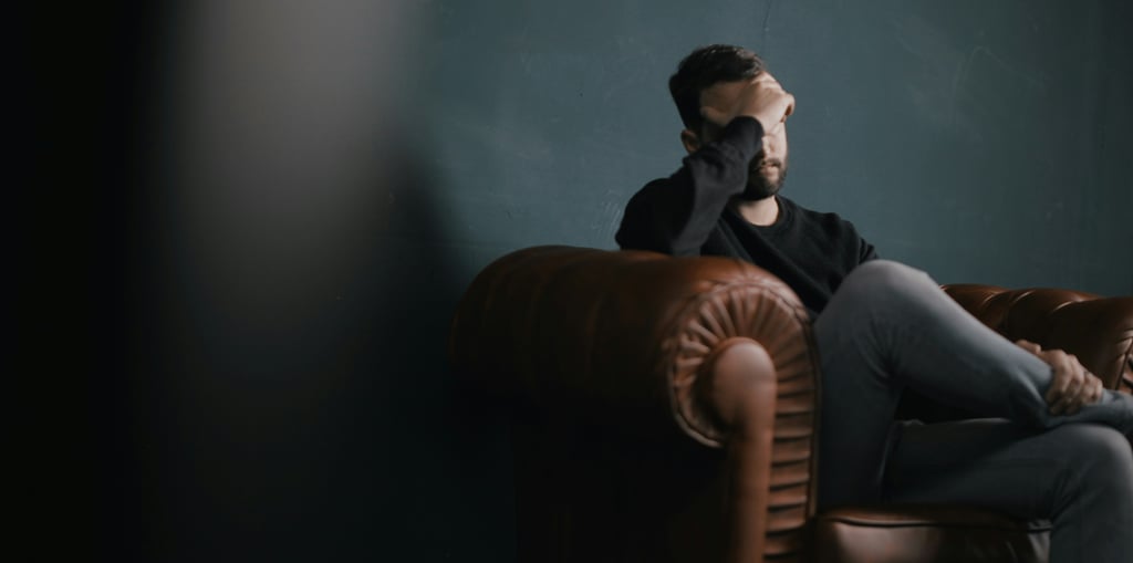 A stressed man sitting on a brown leather armchair holding his head in frustration.