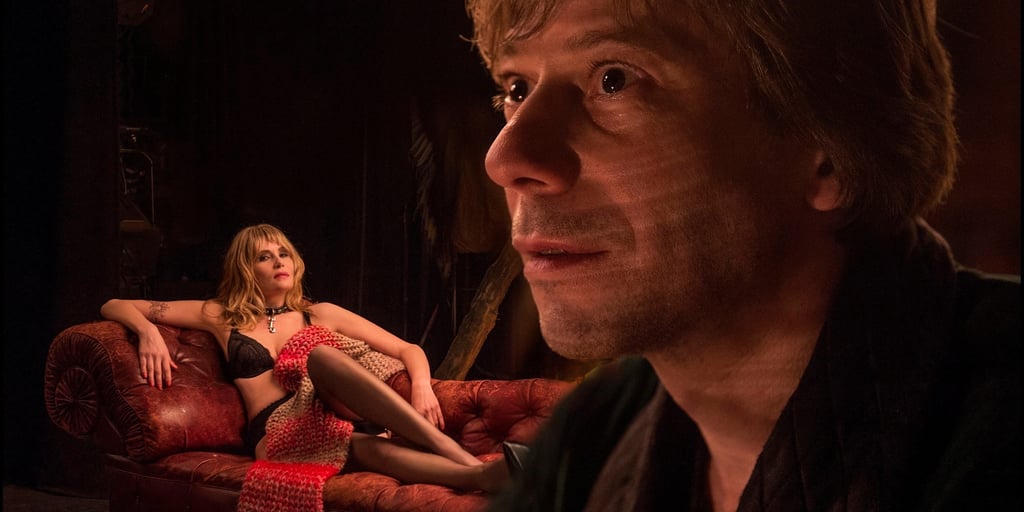 A blond man stares intently while a woman in lingerie poses on a red leather sofa in a dimly lit room.