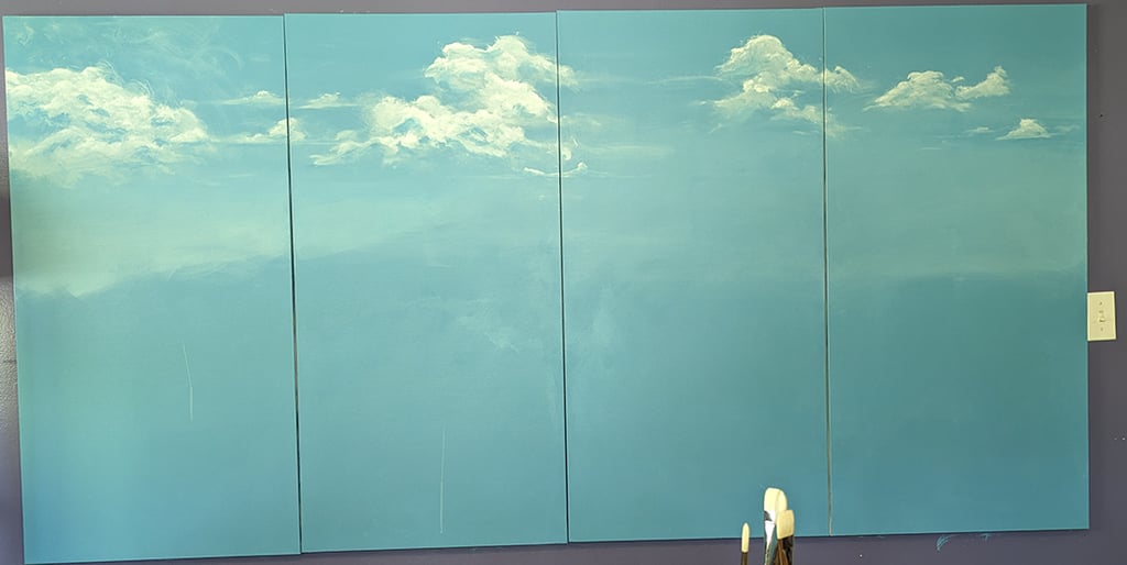 Four stretched canvas panels painted in a seashore blue now with fluffy swirling white clouds painted into the blue.