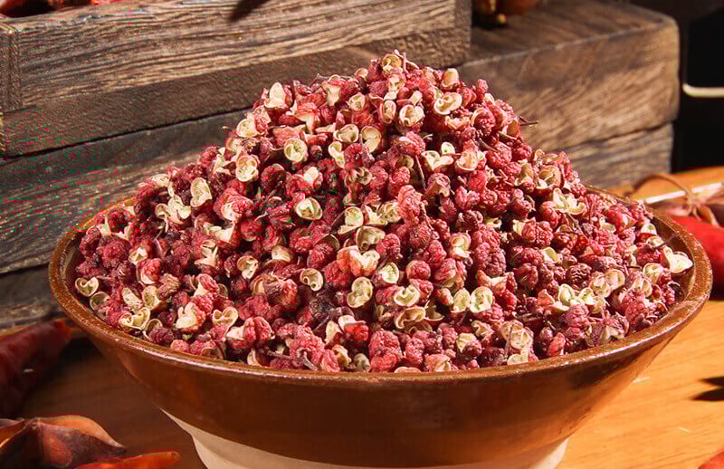 A brown bowl overflowing with red Sichuan peppercorns and dried chili peppers on a rustic wooden table.