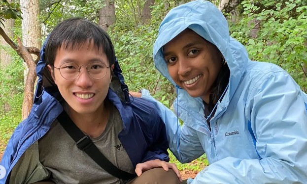 A smiling young man and woman wearing rain jackets while hiking in a lush green forest.