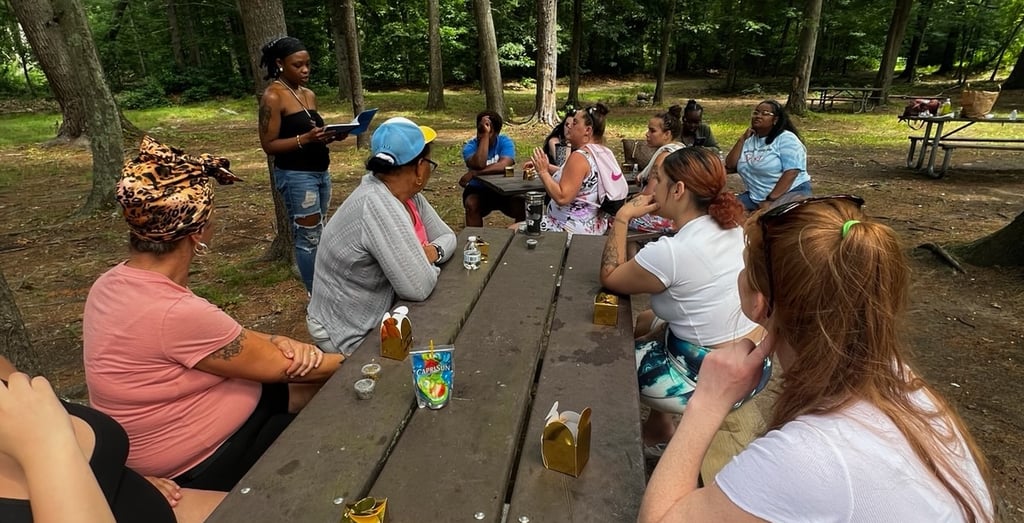 A diverse group of women sitting at a picnic table for an outdoor gathering in a wooded park.