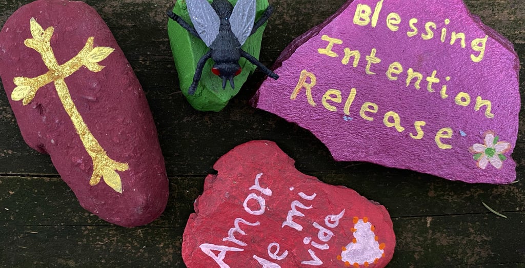 Hand-painted stones with a gold cross, fly figurine, and spiritual messages on a rustic wood background.