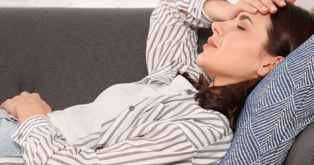 woman laying on a couch with one hand on head and one on stiomach, seems to be in pain