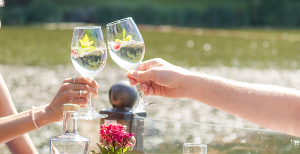 Zwei Hände stoßen mit Weingläsern im sonnigen Restaurant mit Seeblick an, warme Lichtstimmung