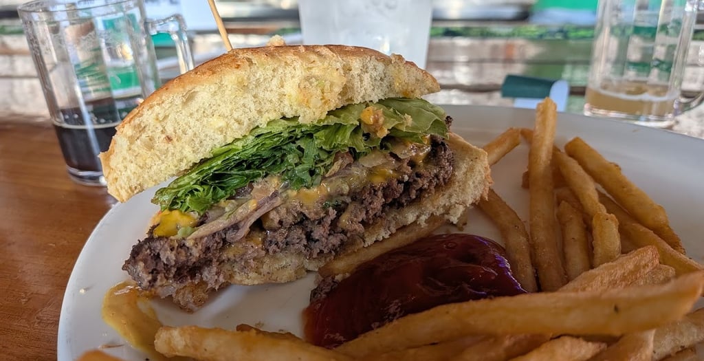 A gourmet cheeseburger cut in half served with crispy french fries and ketchup on a white plate at an outdoor bar.