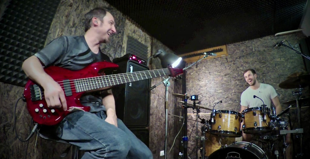 Jozef Bobula on red six-string bass with drummer in a rustic dimly lit rehearsal space.