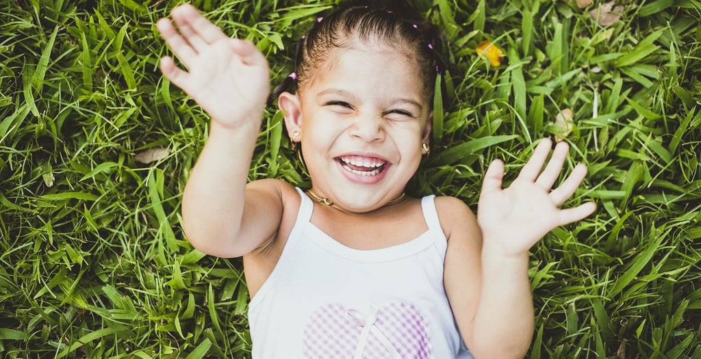 A joyful child in a white heart-patterned shirt lies on grass, smiling broadly with arms raised.