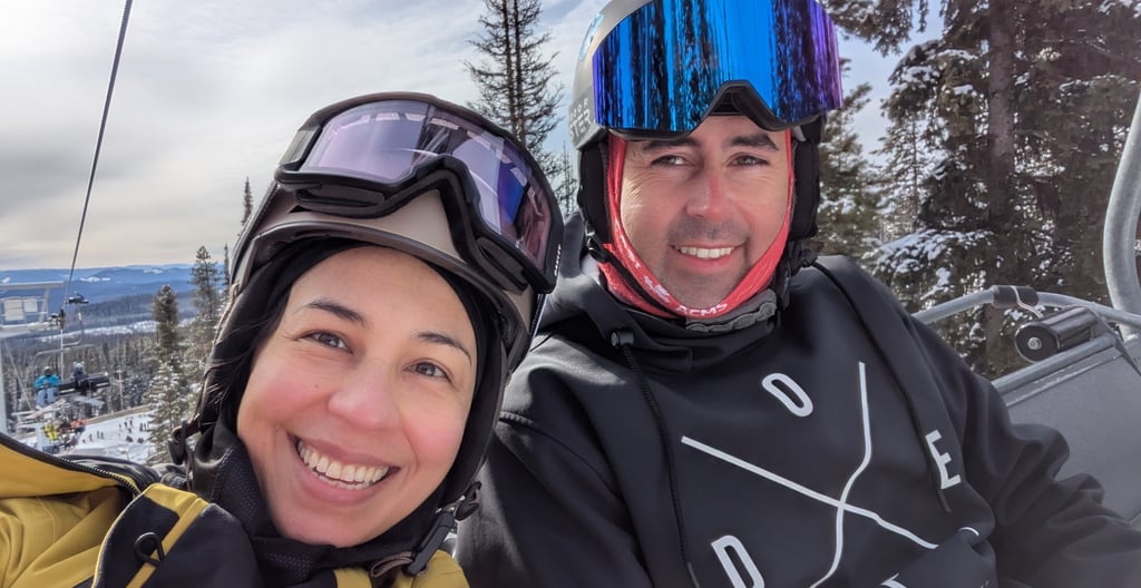 Two skiers smiling on a chairlift with snowy trees and mountains in the background.