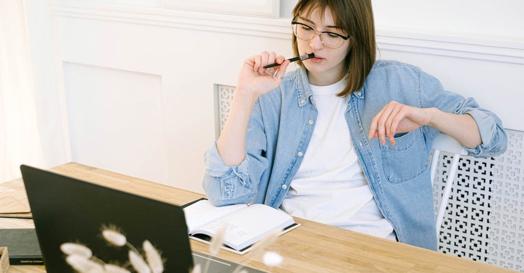 A focused woman wearing glasses and a denim shirt working at a wooden desk with a laptop and notebook.