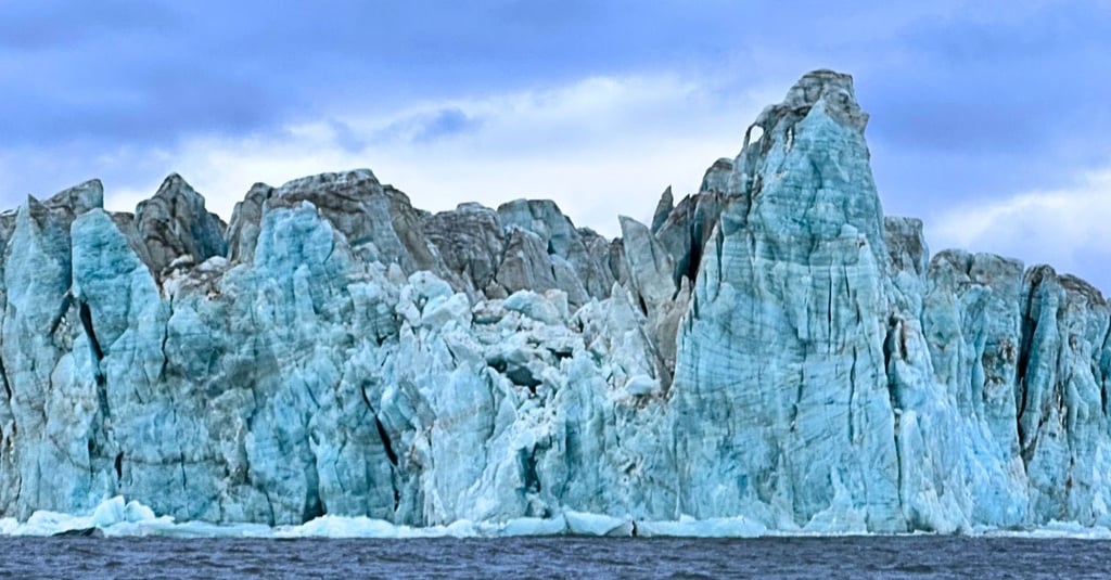 Massive blue glacier ice wall towering over dark ocean water under a cloudy sky.