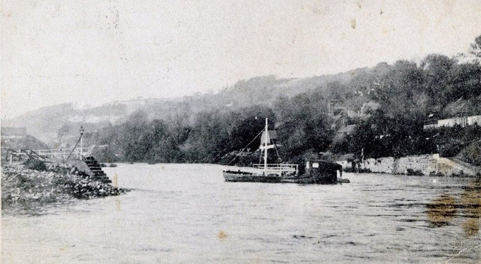 Vintage black and white photograph of a small sailing boat on a river near a wooded shoreline.